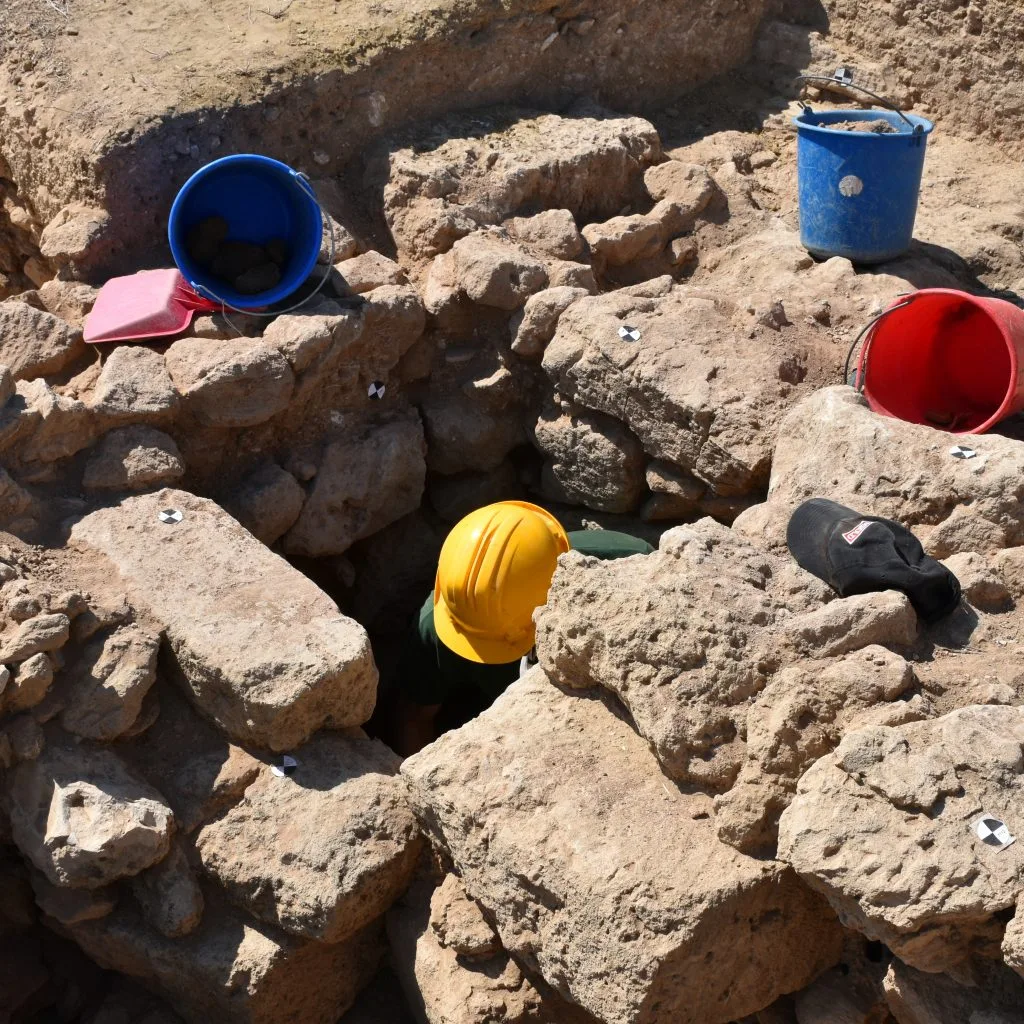 Looking down into a narrow shaft among large rough limestone blocks, with an excavator in a yellow hard hat inside it and buckets on the rocks above