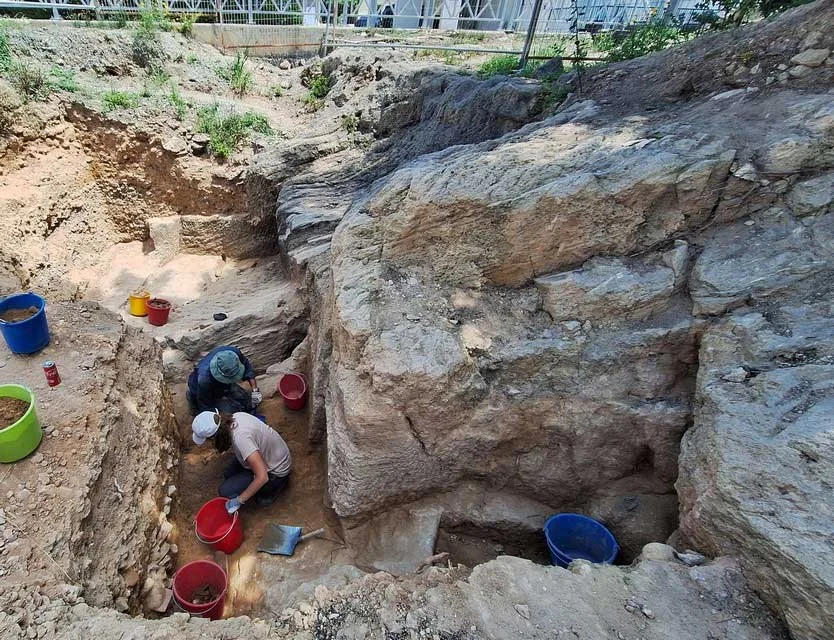 Two excavators in a deep trench, one seated on its rock edge with a notebook and one at the bottom beside a wall of roughly coursed stones