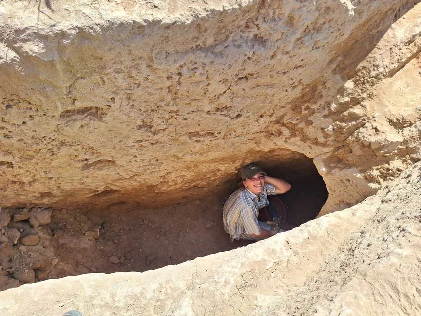 An excavator crouching in a low tunnel mouth at the bottom of a rock-cut shaft, with rubble walling at the base
