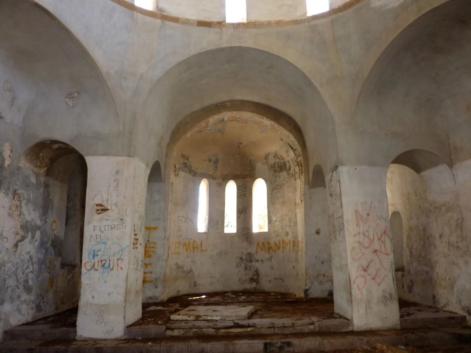 The bare interior of a domed church, its plastered walls and piers covered in spray-painted graffiti, with three narrow arched windows in the apse