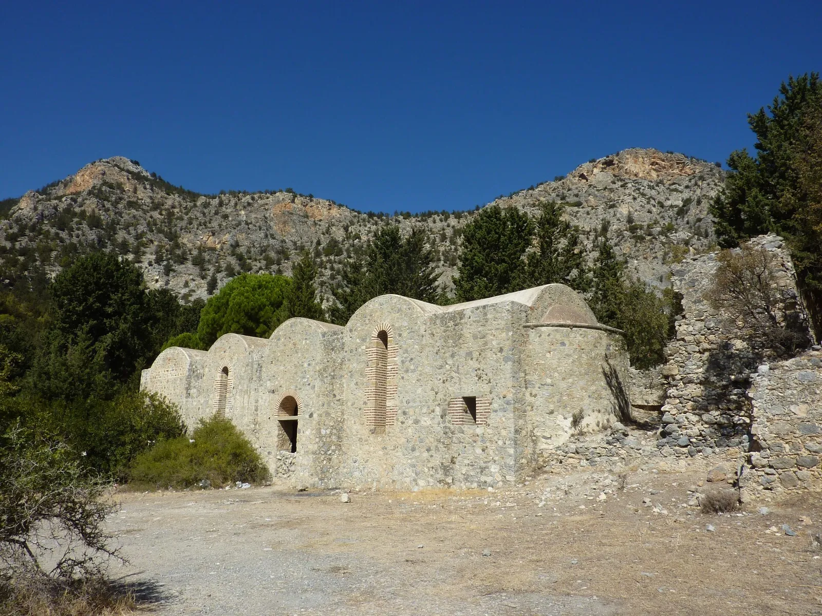 A long low stone building with a row of arched recesses along its wall and a small window, roofless ruins to its right and a rocky mountain ridge behind