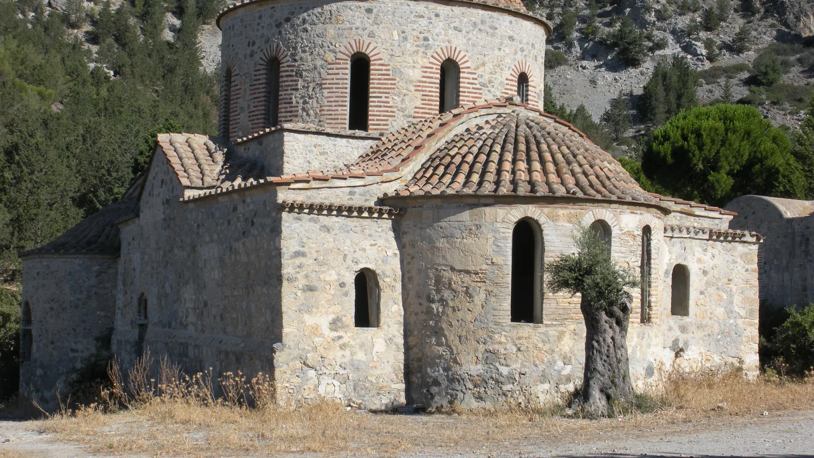 A domed stone church with tiled roofs and a rounded apse, standing on bare ground below a steep wooded mountainside