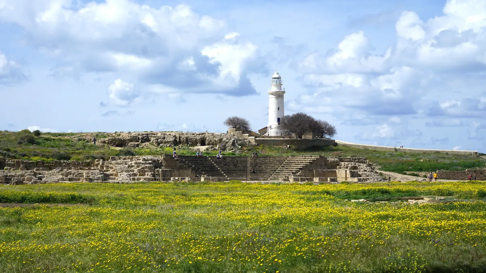 A white lighthouse tower on a low rocky hill with low stone buildings and trees at its foot, above the stone seating of a small ancient theatre and a field of yellow flowers