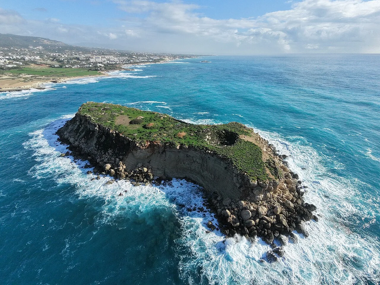 Aerial view of a small flat-topped rocky islet with sheer cliffs in turquoise sea, white surf along its edges and a coastline with buildings behind