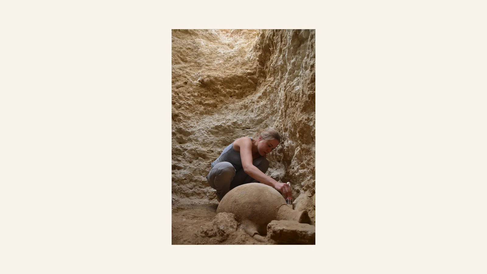 An excavator crouches in a deep trench cut into pale rock, brushing the shoulder of a large clay storage jar lying on its side in the earth