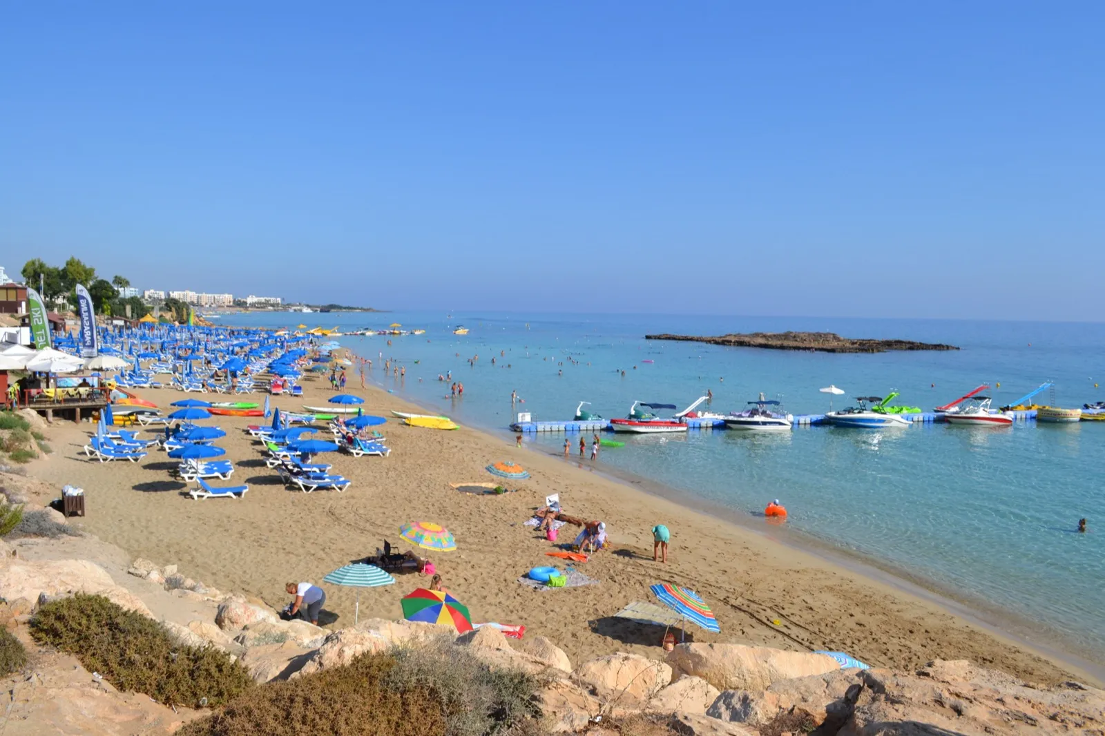 A sandy beach crowded with blue umbrellas and sunbeds curving round shallow turquoise water, with moored boats and a low rocky islet offshore