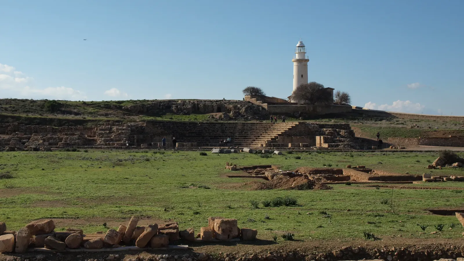 A grassy open square edged with low stone foundations and excavation trenches, with the curved stone seating of a small theatre and a white lighthouse on the hill beyond