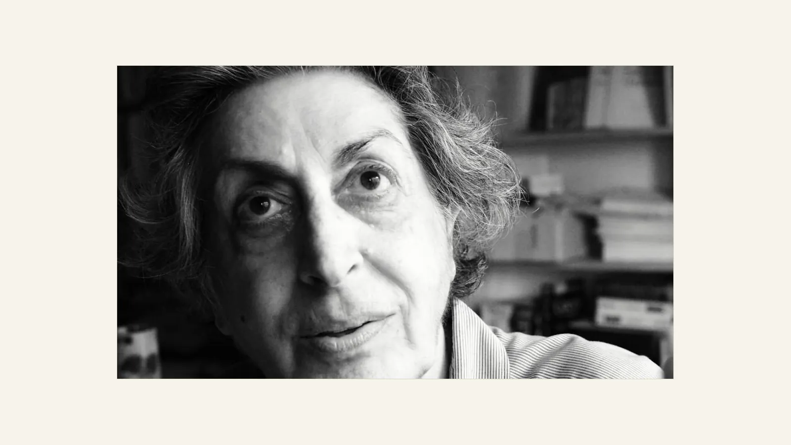 Black-and-white close portrait of a grey-haired woman in a striped shirt, looking at the camera, with shelves of books behind her