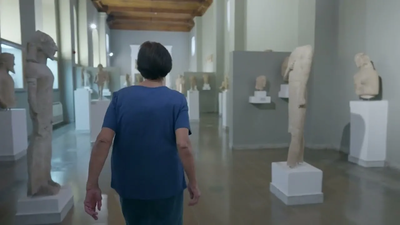 A woman seen from behind walks down a museum gallery lined with ancient limestone statues on white plinths, under a wooden ceiling