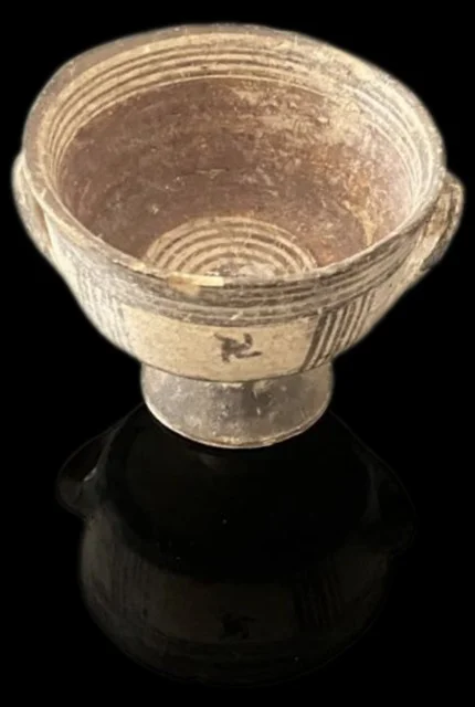 A small footed bowl with a loop handle on each side, painted in dark bands with concentric rings inside and a hooked-cross motif in a panel on the outside, photographed on a black ground
