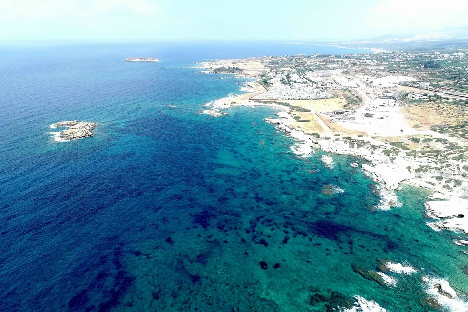 Aerial view of a rocky limestone coastline with turquoise shallows, a small rocky islet close to shore, a second islet further out, and low buildings along the coast
