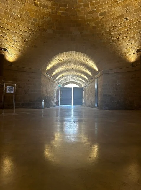 Long vaulted stone passage lit by warm uplights along its walls, with a polished floor and tall doors at the far end