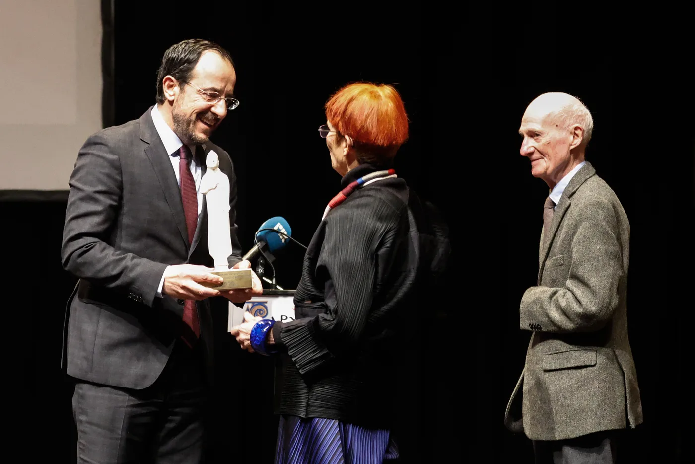 On a dark stage, a man in a grey suit hands a small white statuette to a woman with red hair while an elderly man in a tweed jacket stands beside her