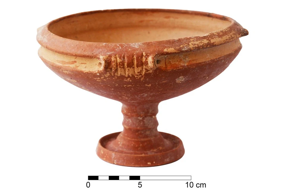 Red-brown clay bowl on a tall stemmed foot with a flat base, a raised band and small lugs below the rim, beside a 10 cm scale bar