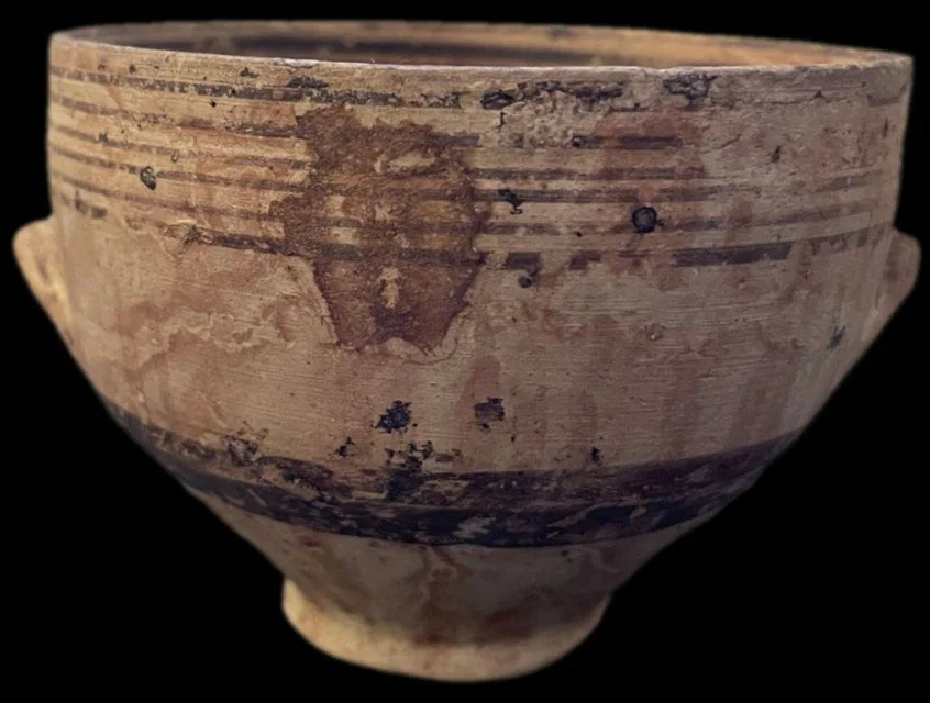 A pale clay bowl on a short foot with a stub handle on each side, painted with thin dark bands round the rim and a broad dark band lower down, against a black background
