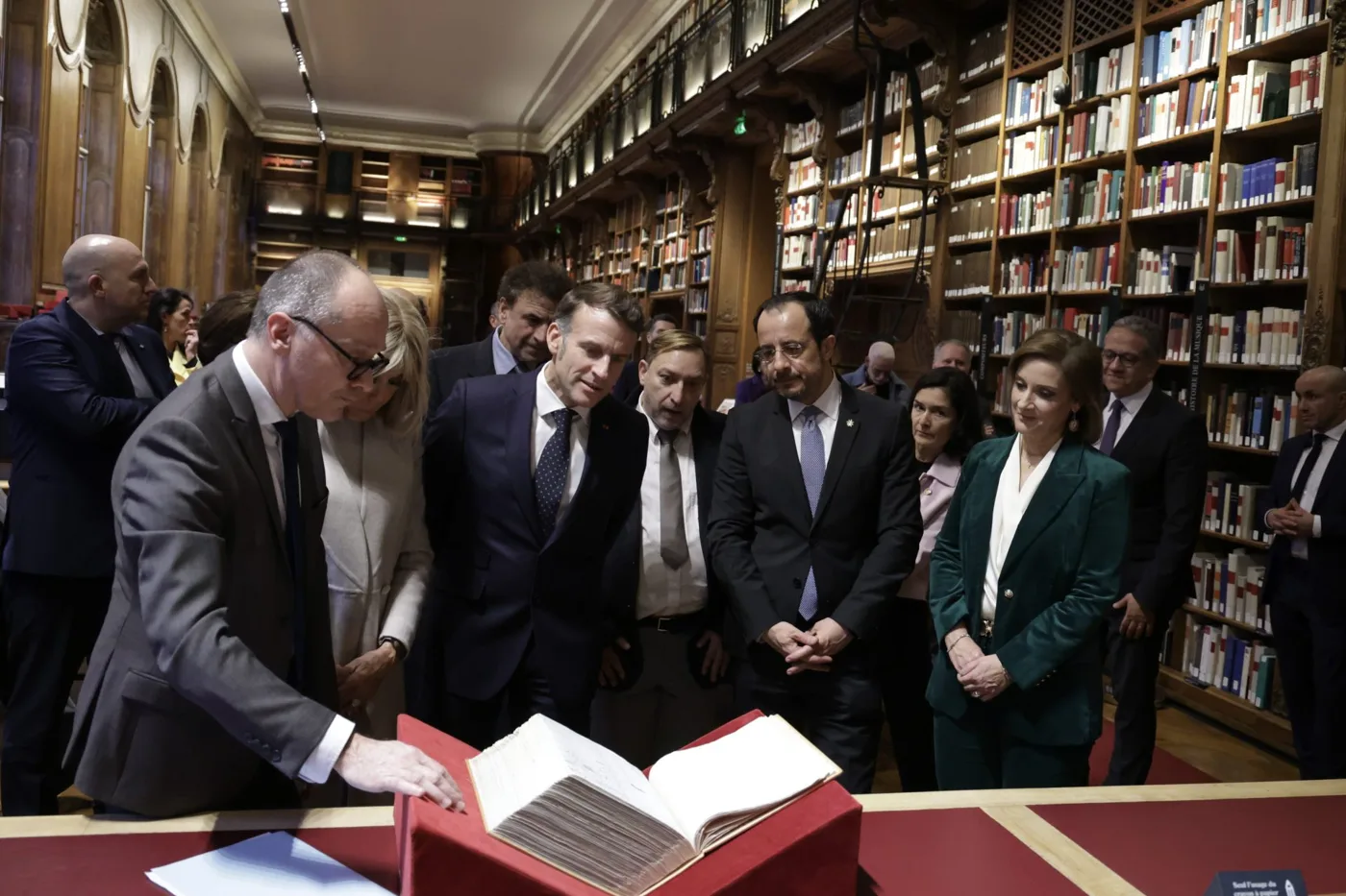 A man in a dark suit leans over a large open manuscript on a red cushion in a library lined with bookshelves, watched by a group of officials