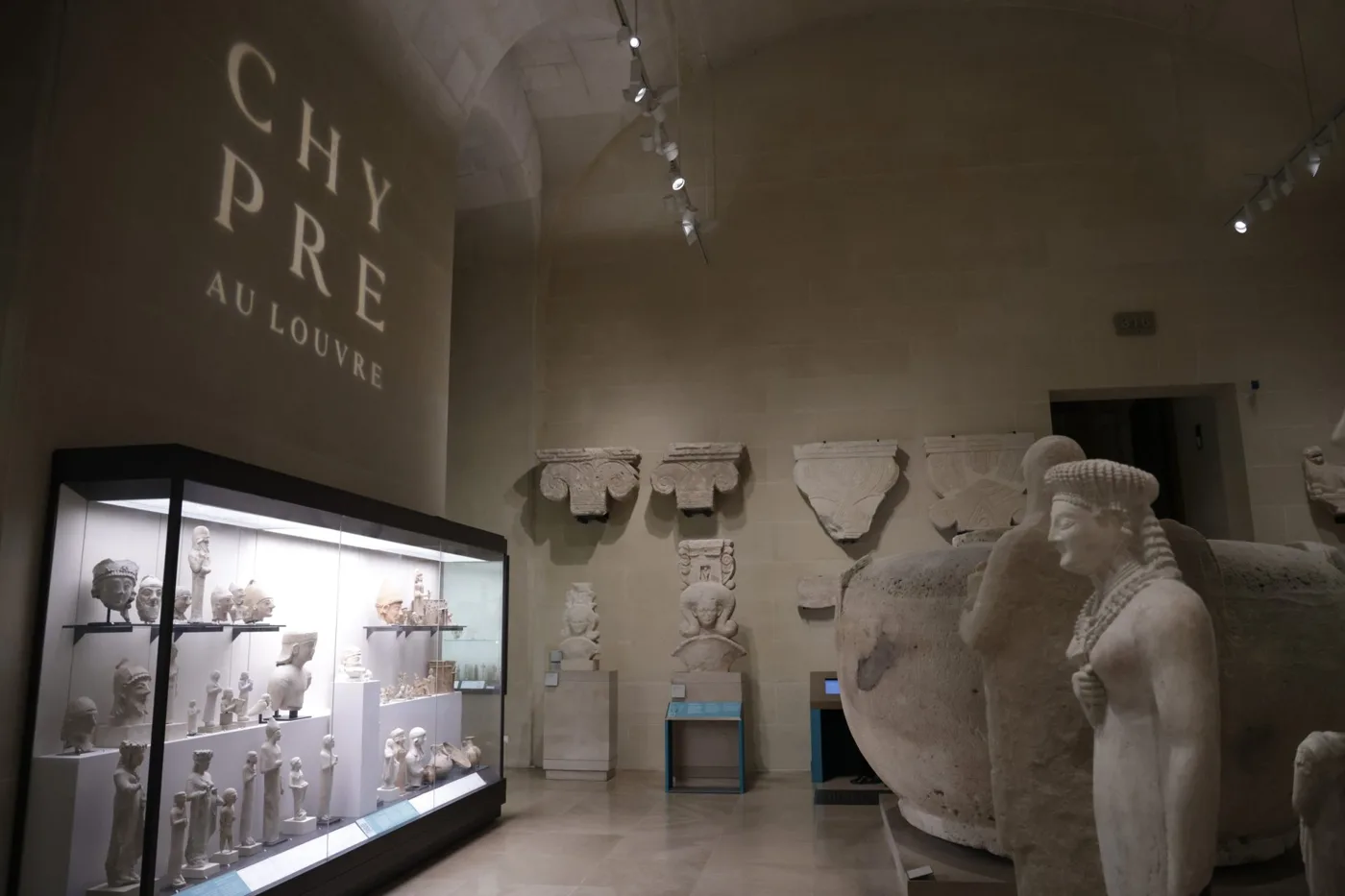 Museum gallery with a vaulted ceiling and limestone statues, capitals and a large stone vase, a lit wall case of small figures, and the words Chypre au Louvre projected on the end wall