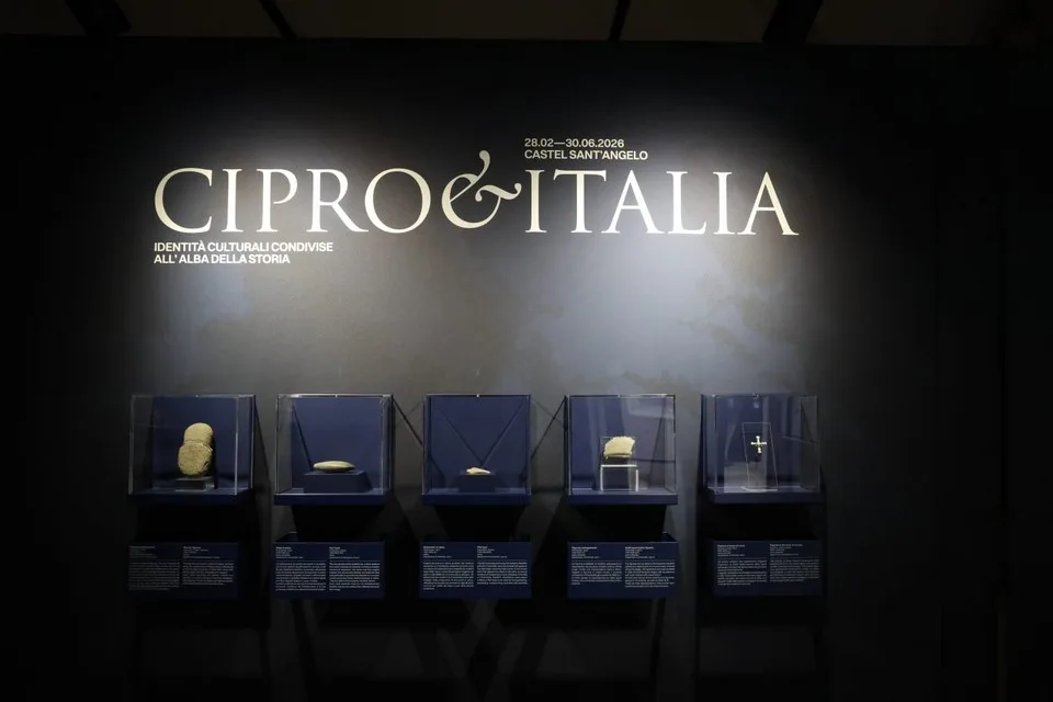 Dark exhibition wall with the title Cipro e Italia in large pale letters above a row of five lit glass cases, each holding a small stone or metal object, with labels below