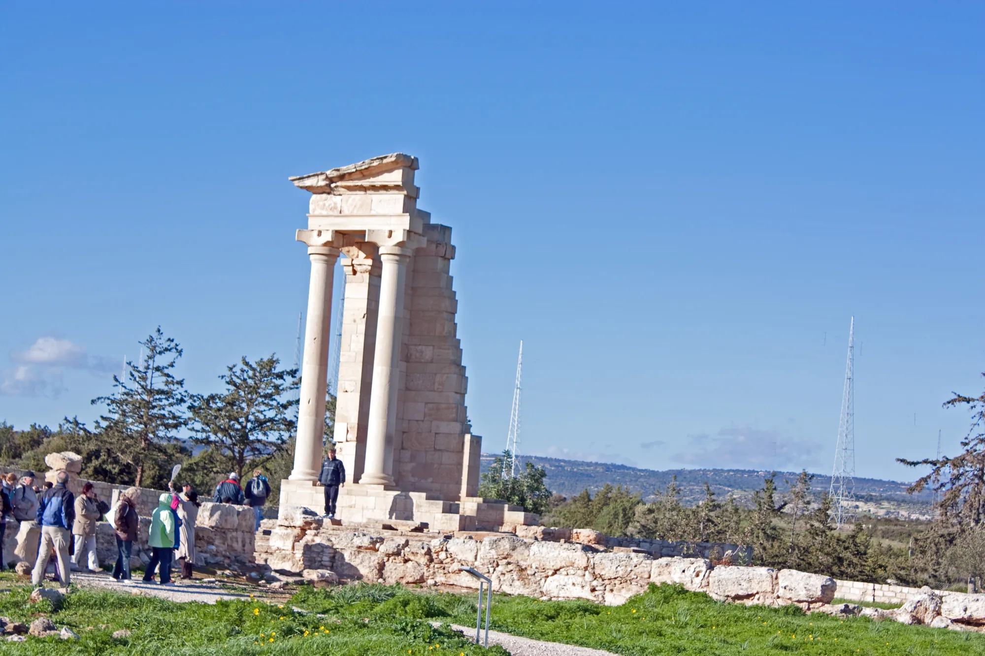 Stone columns of a small temple standing on a stepped platform under a blue sky, with visitors walking among the ruins