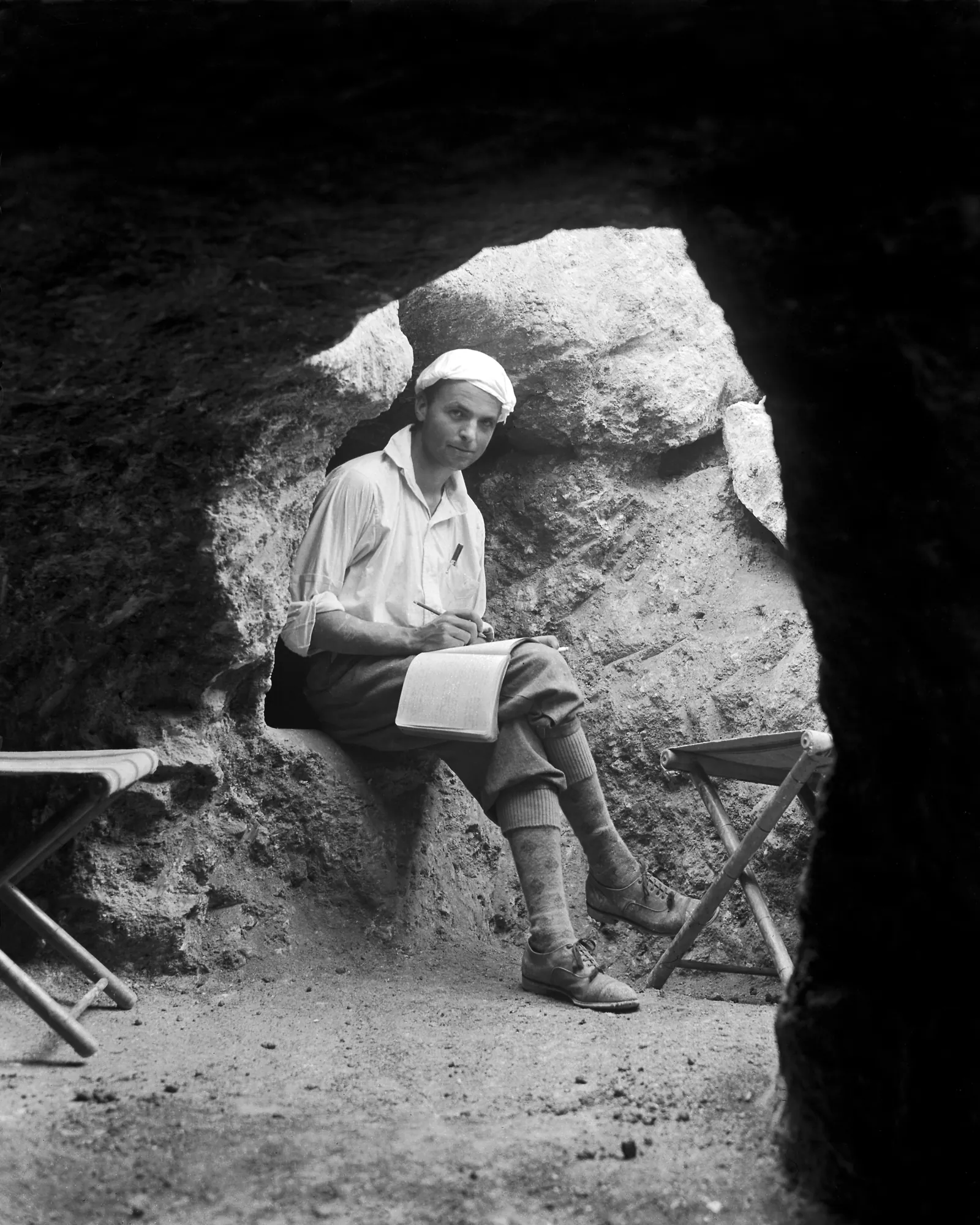 Black-and-white photograph of a man seated in the dark entrance of a rock-cut tomb, writing in a notebook