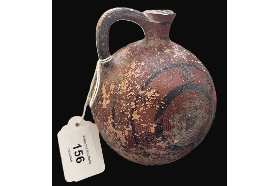 A round-bodied red clay jug with one handle and a broken rim, painted with black concentric circles and bands, with a paper auction tag numbered 156 tied to the handle, on a black ground