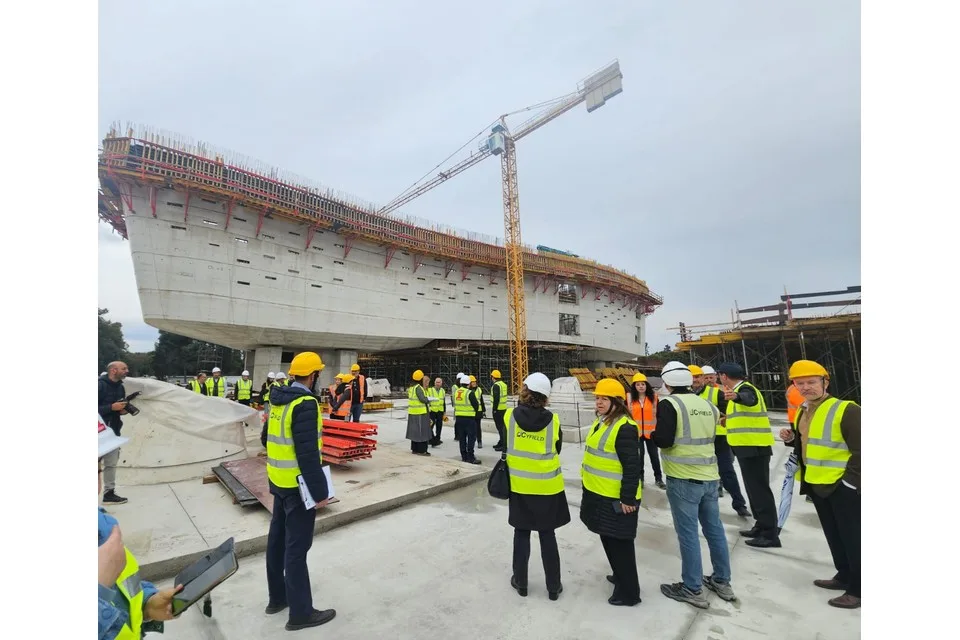 Visitors in hard hats and yellow high-visibility vests stand on a construction site in front of a long curved concrete building with a crane above it
