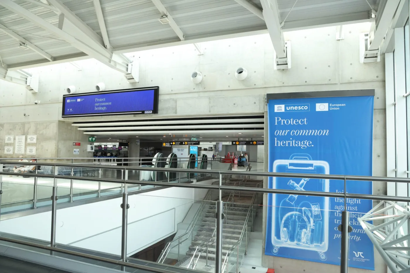 A tall blue poster with an X-ray-style drawing of a suitcase and the words Protect our common heritage, beside the stairs of an airport departures hall, under UNESCO and European Union logos