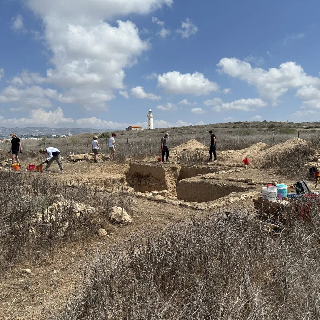 Excavators with buckets working round a rectangular trench cut into dry ground, with low stone walls at its edge and a white lighthouse on the skyline behind