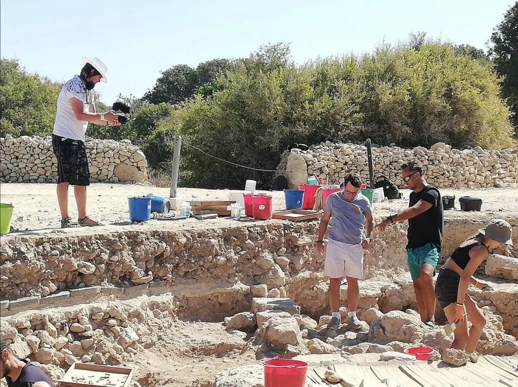Excavators working among rough stone foundations in a trench, with a man filming from the edge above, buckets and finds trays along the rim and dry-stone walls behind