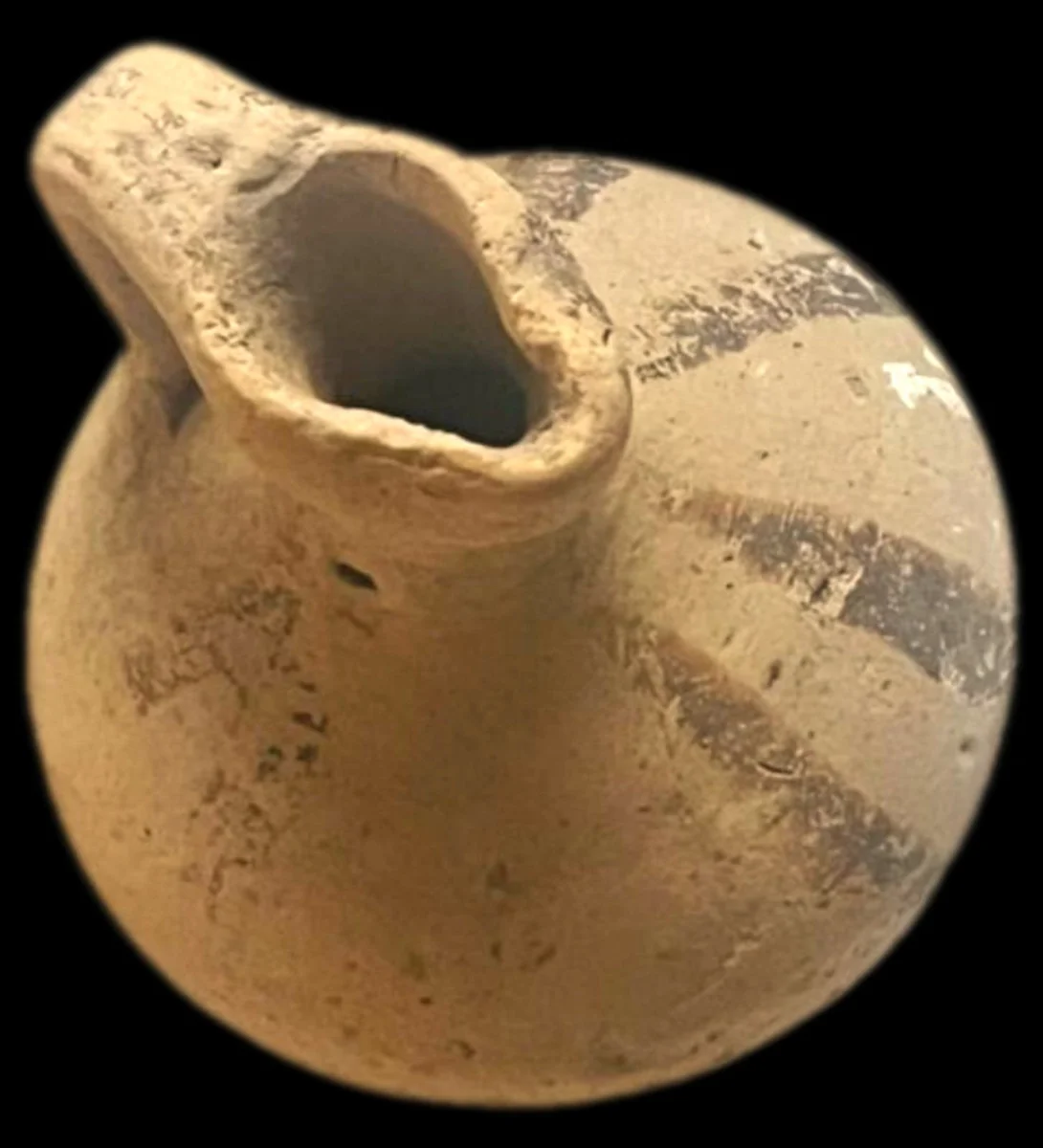 Small round-bodied clay juglet seen from above, with a pinched mouth, pale buff surface and dark painted bands on the body, against a black background