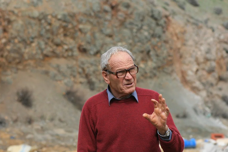 An elderly man with grey hair and dark-rimmed glasses, in a red jumper over a checked shirt, speaking and gesturing with one hand in front of a scrub-covered rocky slope