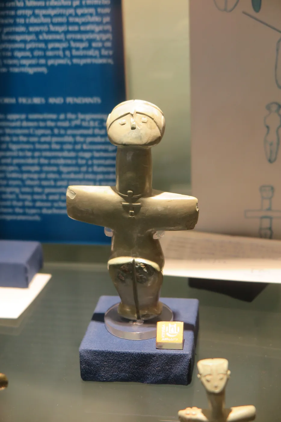 A pale green stone figure with outstretched arms, bent legs and a carved face, wearing a tiny figure of the same cross shape on its chest, standing in a museum case