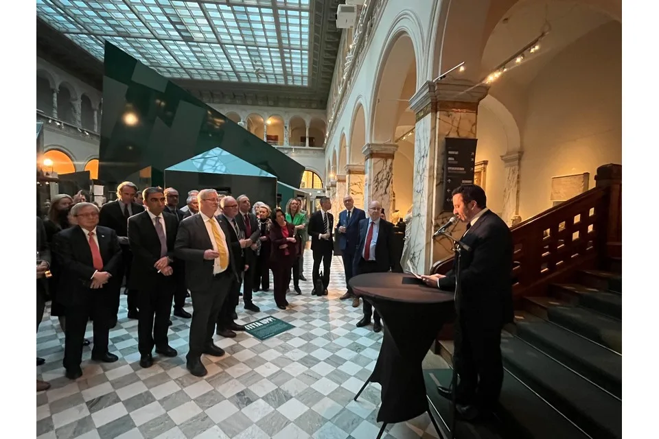 A man in a dark suit speaks at a lectern beside a staircase to a standing audience in formal dress, in a museum hall with marble columns, a chequered floor and a glass roof