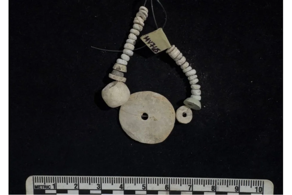 A loop of small white disc beads threaded on a wire with a larger round bead and a flat perforated disc, on a black background above a 10 cm scale, with a paper label reading MV765