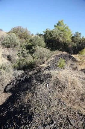 A long low mound of black slag partly covered by dry grass and shrubs on a hillside, under a clear blue sky, with a small scale marker in the middle