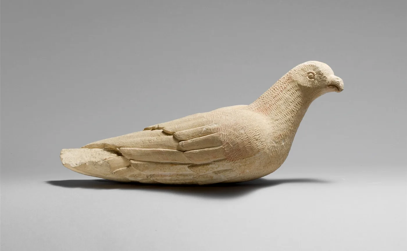 A pale limestone statuette of a resting bird with folded, incised wings and a small hooked beak, on a grey ground
