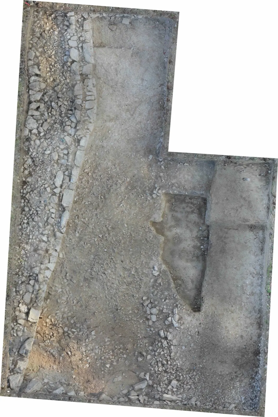 Overhead photograph of an L-shaped excavation trench, with a broad band of packed rough stones running down its left side and bare earth floors to the right