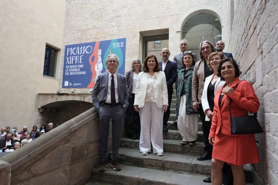 A group of people standing on the stone steps of a courtyard beneath a banner reading Picasso Xipre, Trobades amb la ceramica del Mediterrani, 06-06-26 to 27-09-26
