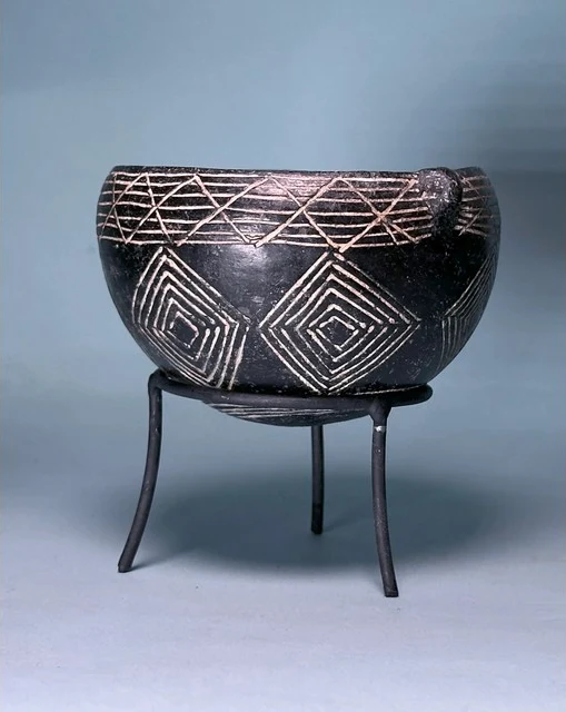 A round-bottomed black bowl with a small lug at the rim, decorated with white-filled incised bands of crossing lines and nested lozenges, resting on a black metal tripod stand against a blue-grey ground