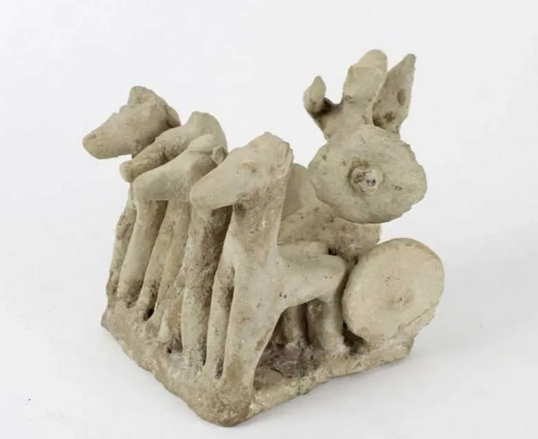 Pale clay model of four horses side by side drawing a box in which two figures stand, one holding a round shield, with a round wheel at the side, on a white background