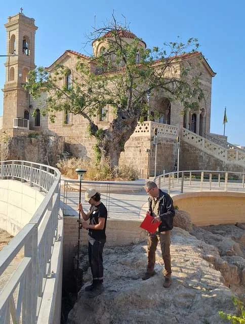 Two men on bare bedrock beside a modern railed walkway, one holding a surveying pole and the other a red folder, below a stone church with a bell tower and a tree in front