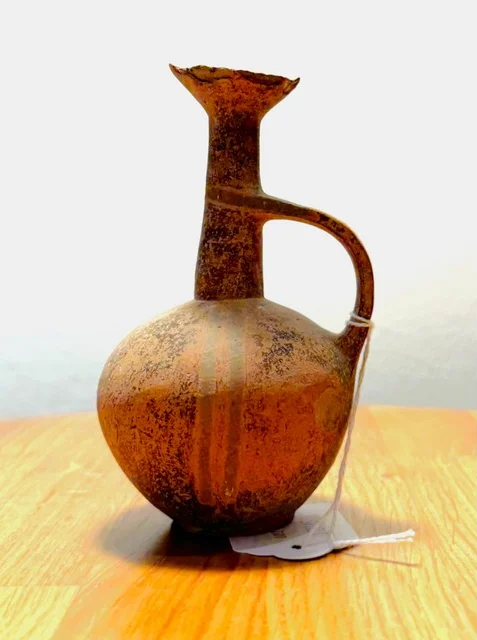 A small round-bodied reddish-brown jug with a tall narrow neck, flaring mouth and a looped handle, standing on a wooden table with a paper tag tied to the handle