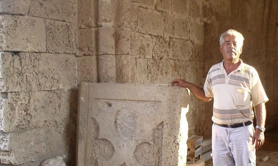 A grey-haired man in a striped shirt resting his hand on a large square stone slab with a partly carved relief, standing against a wall of cut stone blocks