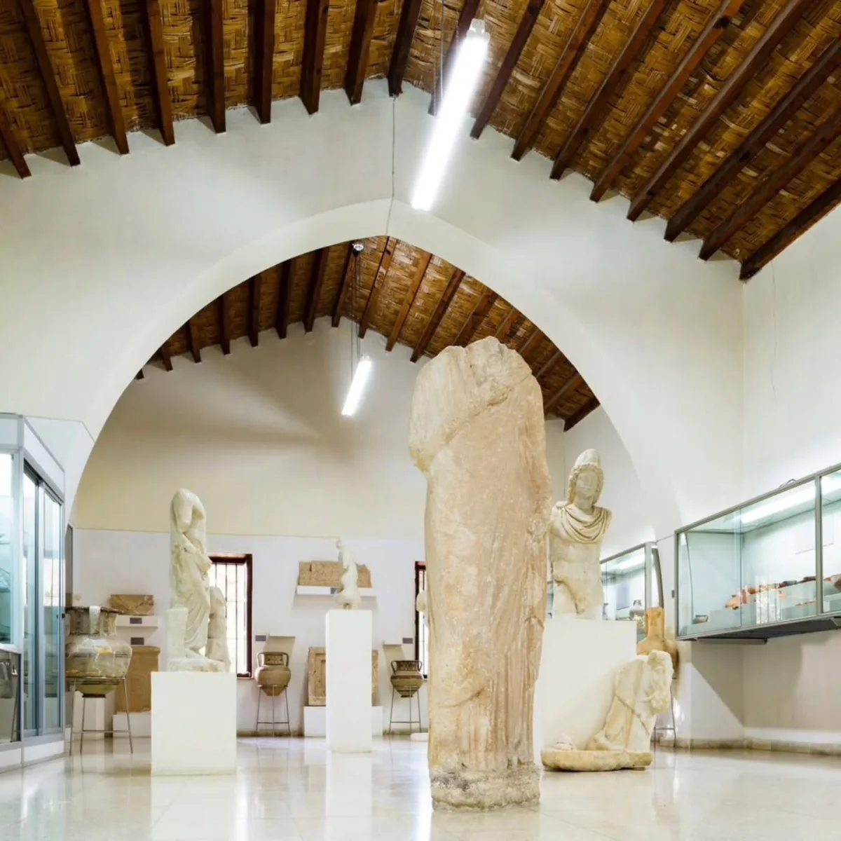 A white-walled museum hall under a pointed stone arch and a timber and reed ceiling, with a headless draped marble statue in the foreground, other statues on plinths and pottery in glass cases