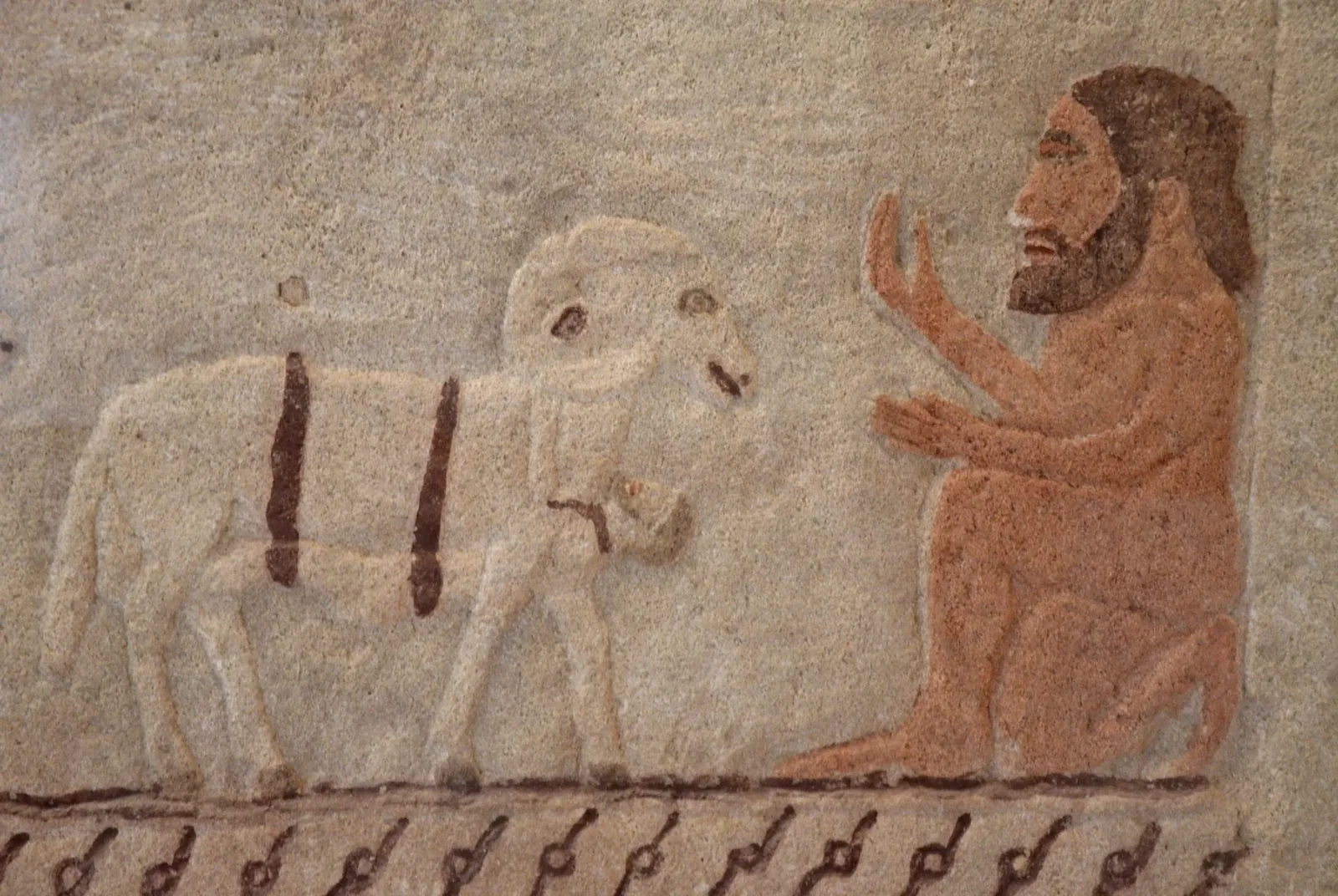 Painted relief on pale limestone of a seated, bearded, naked man with raised hands facing a ram with a small figure beneath it, above a band of spiral pattern