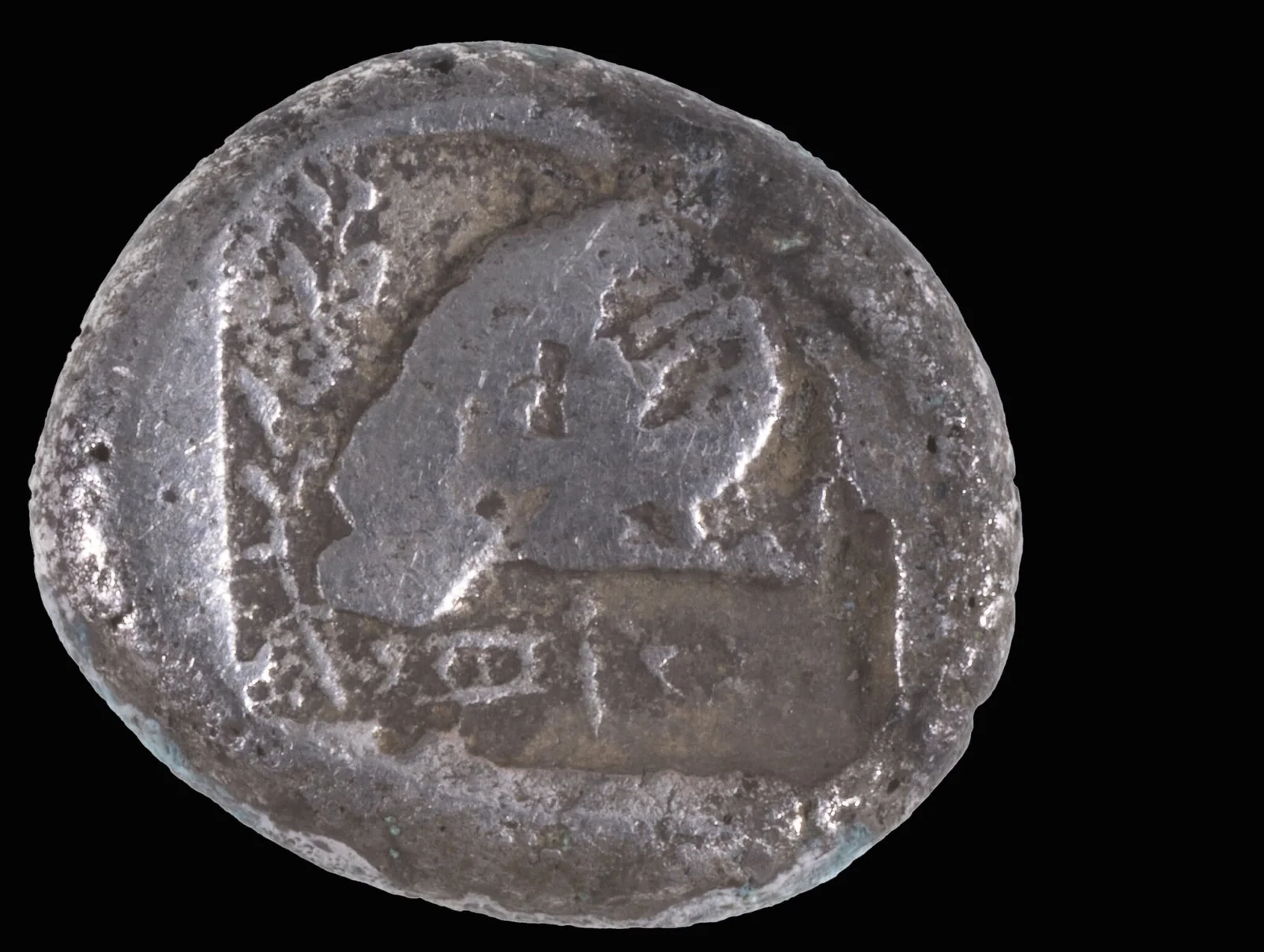 A worn grey silver coin on a black ground, showing a ram's head inside a sunken square, with a branch on the left and faint signs along the bottom