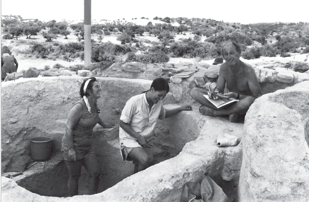 Black-and-white photograph of three people in a stone-lined excavation pit on a scrubby hillside, a man seated on the rim writing in a notebook and two women standing in the pit below him