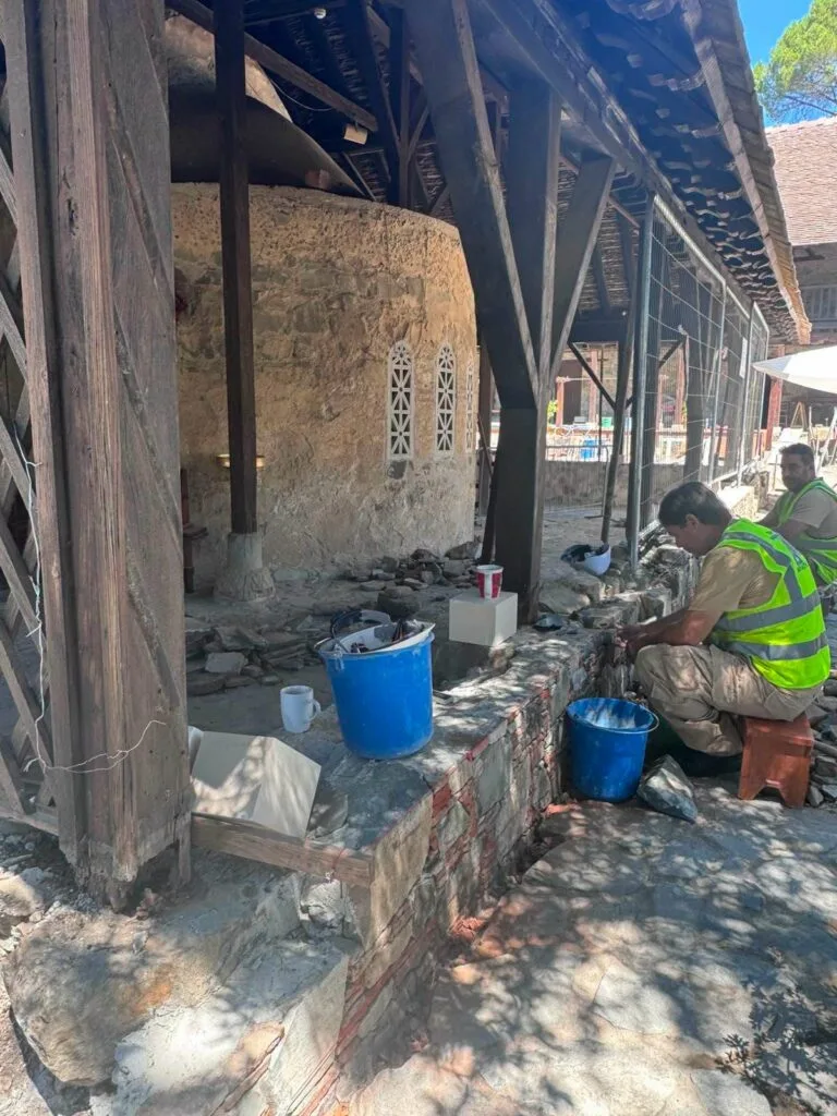 Conservators in high-visibility vests working on a low stone wall under the timber outer roof of a church, beside the curved stone apse, with buckets and tools on the wall