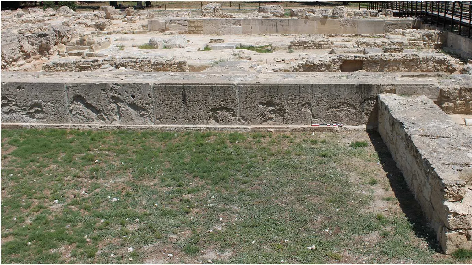 A long wall of large weathered limestone slabs with faint carved marks, a scale bar at its foot and a grassy strip in front, with excavated stone foundations behind