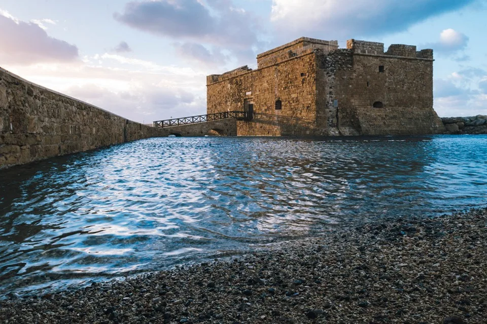 A square stone fort with crenellated walls standing at the end of a harbour mole, reached by a footbridge, with calm water and a pebble shore in front